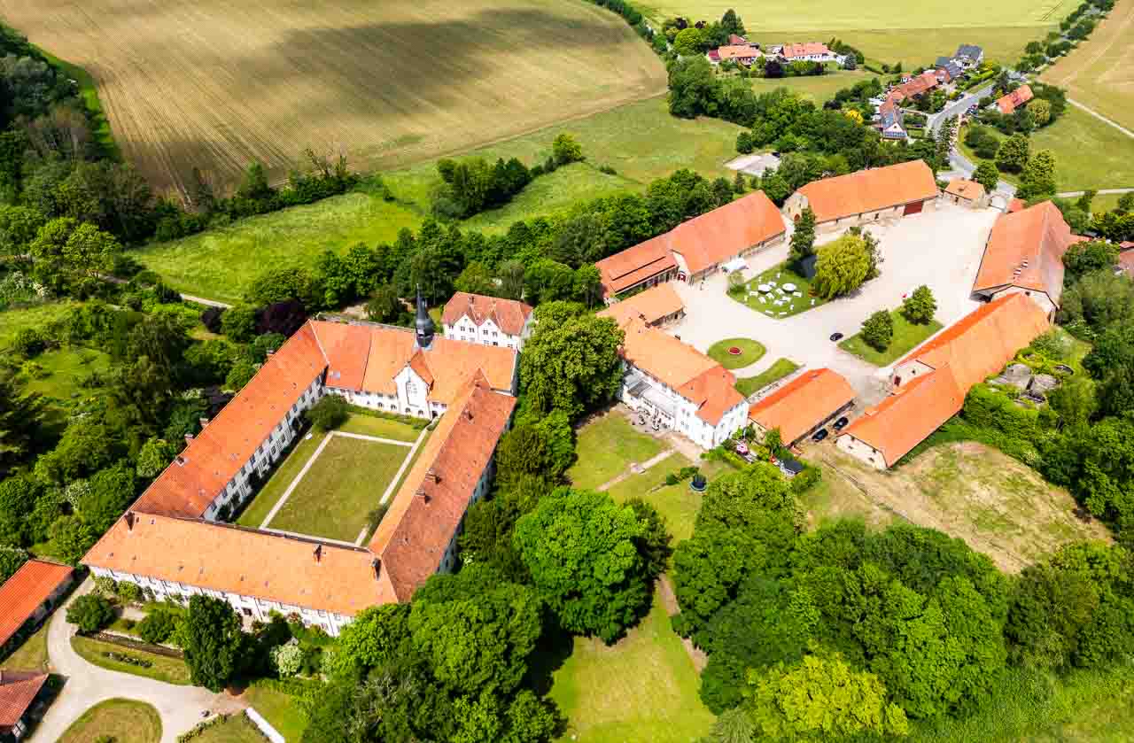 Luftaufnahme eines Schlosses mit rotem Dach, umgeben von gruenen Baeumen und Feldern. Die Architektur versprueht einen historischen Charme.