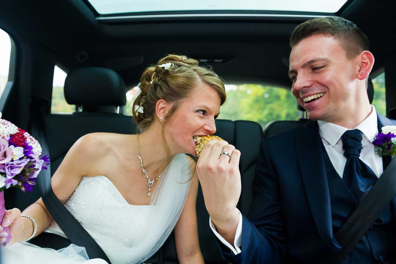 Entzueckende Braut, die im Auto von ihrem Braeutigam einen Snack bekommt. Sie strahlt in ihrem weissen Kleid, er elegant im dunklen Anzug. Liebe!
