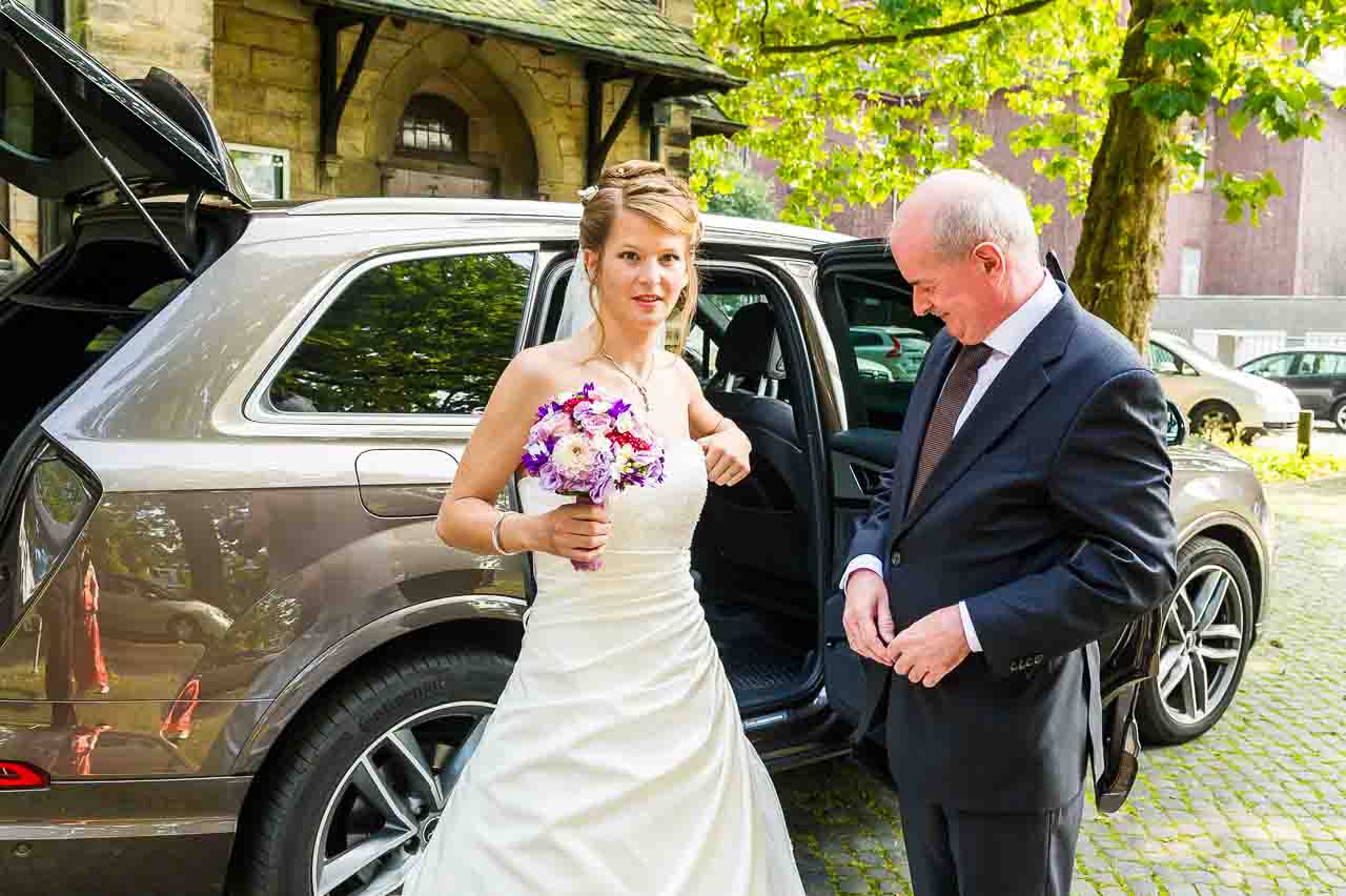 Eine strahlende Braut mit Blumenstrauss steigt aus einem eleganten Auto, begleitet von ihrem Vater. Ein schoener Moment vor dem Standesamt.