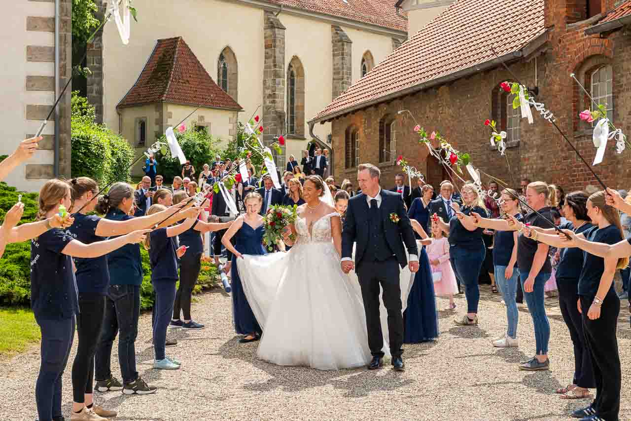 Strahlende Braut mit Braeutigam nach der kirchlichen Trauung. Spalier mit Rosenstaeben und weissen Baendern fuer das glueckliche Paar.