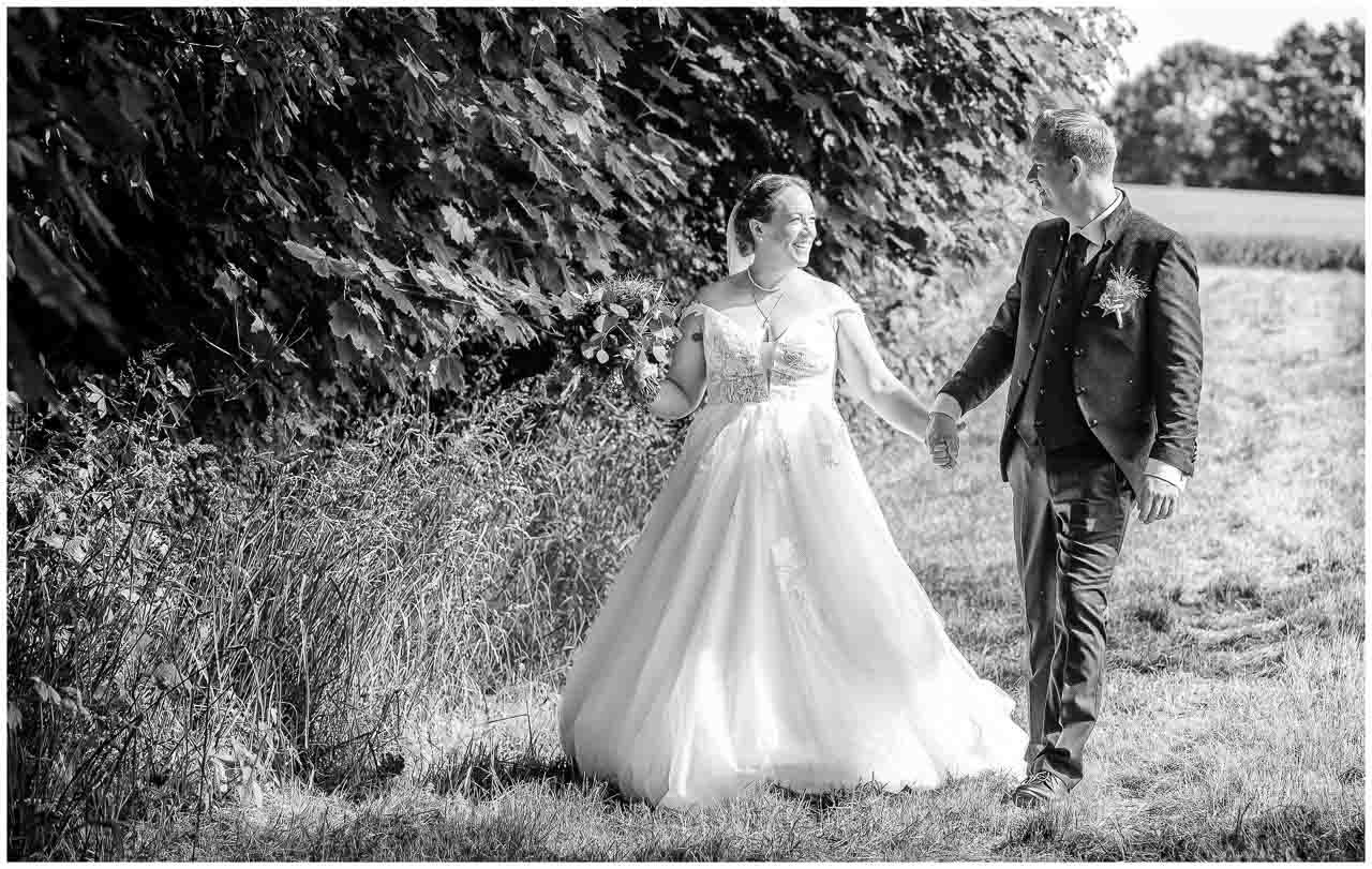 Schwarz-Weiss-Foto: Braut im eleganten Brautkleid und Braeutigam Hand in Hand. Ein zaertlicher Moment in der Natur, voller Liebe und Zuneigung.