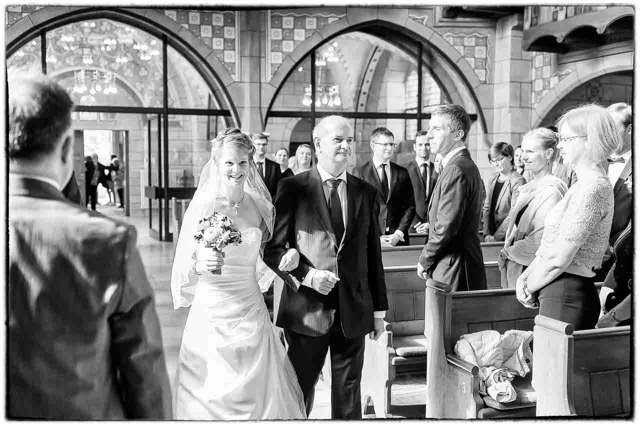 Wunderschoene Braut wird in der Kirche von ihrem Vater zum Altar geleitet. Die Gaeste beobachten das zauberhafte Brautpaar andachtsvoll.