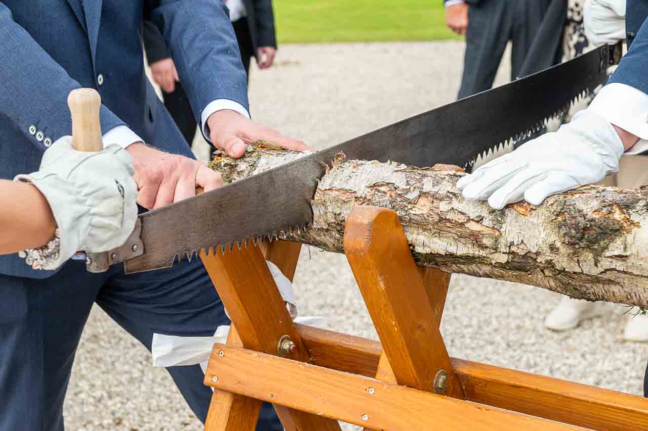 Brautpaar saegt Baumstamm bei Hochzeit. Traditioneller Brauch mit Handsaege und Holzsaegebock. Symbol fuer Zusammenhalt und gemeinsame Zukunft.
