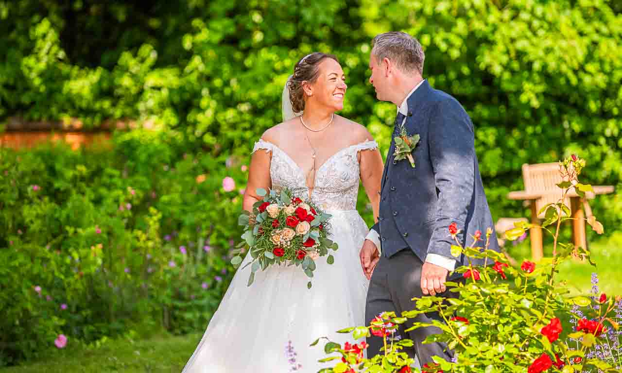 Brautpaar im Garten: Die Braut im eleganten Kleid mit Blumenstrauss, der Braeutigam im Anzug. Romantische Hochzeit in den Gaerten.