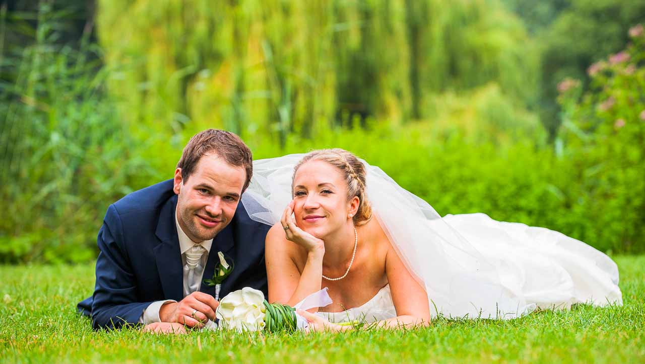 Ein strahlendes Brautpaar, verliebt im Gruenen liegend, geniesst die Atmosphaere im Park. Die Braut mit ihrem schoenen Kleid und der Braeutigam im Anzug.