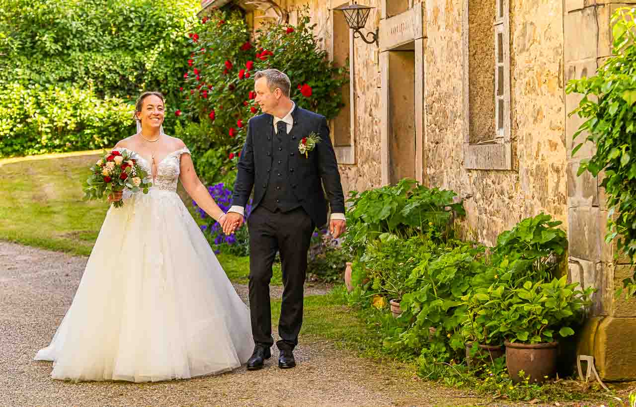 Brautpaar haelt Haende im Park. Die Braut im elfenbeinfarbenen Kleid mit Blumenstrauss, der Braeutigam im eleganten Anzug. Romantische Hochzeit.