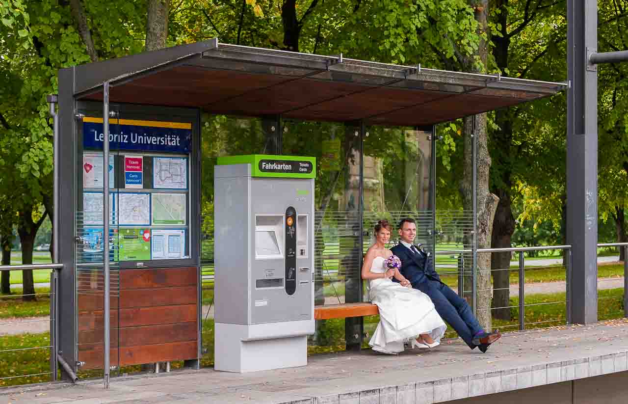Ein stilvolles Brautpaar geniesst den Moment in den Herrenhaeuser Gaerten, sitzend an einer Haltestelle. Die elegante Braut strahlt neben ihrem Braeutigam.
