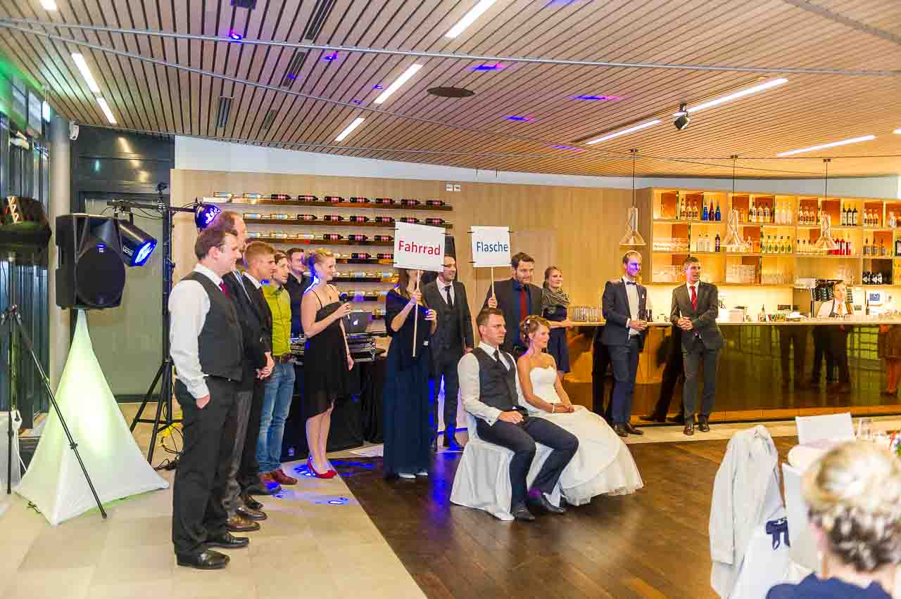 Stilvolles Brautpaar sitzt bei der Hochzeitsfeier. Freunde halten Schilder mit 'Fahrrad' und 'Flasche'. Eine liebevolle Szene in festlichem Ambiente.