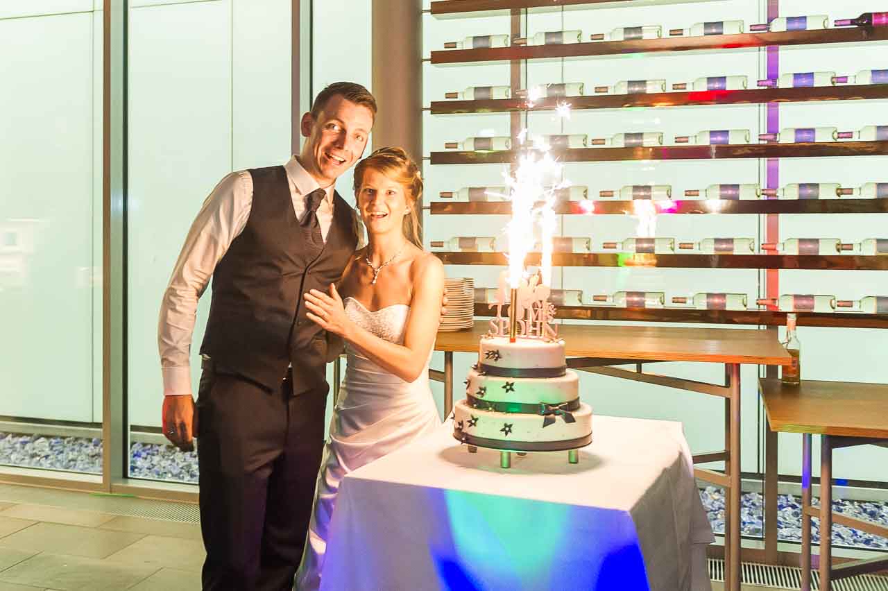 Ein strahlendes Brautpaar vor einer modernen Location, mit einem festlichen Hochzeitstorte, dekoriert mit Sternen und spruehenden Fontaenen, fuer den schoenen Tag.