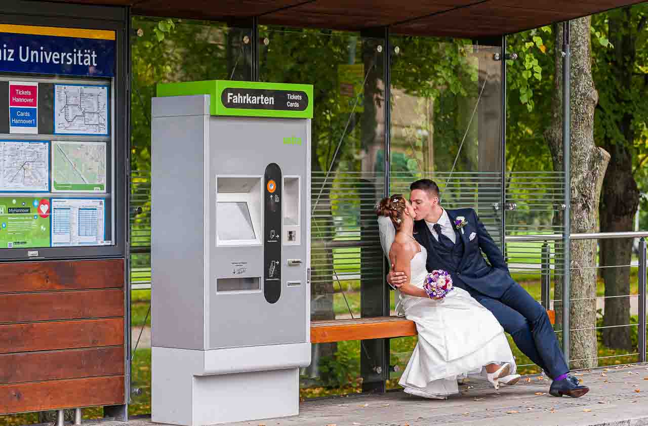 Ein verliebter Braeutigam kuesst seine Braut an einer Bushaltestelle in Hannover. Das Paar sitzt auf einer Bank neben einem Fahrkartenautomaten.