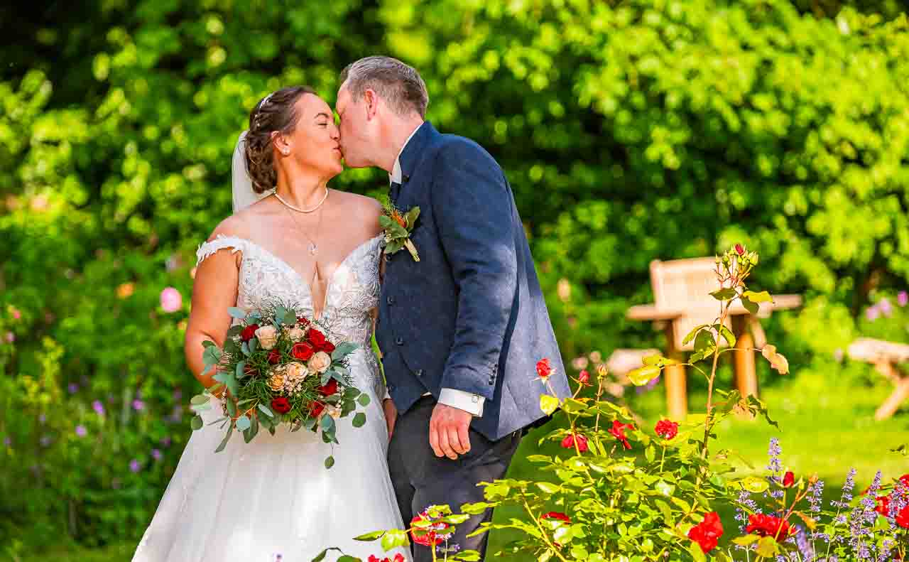 Romantischer Kuss im Garten: Brautpaar teilt innigen Moment. Die Braut im eleganten Kleid mit Blumenstrauss, der Braeutigam im Anzug. Liebe liegt in der Luft.