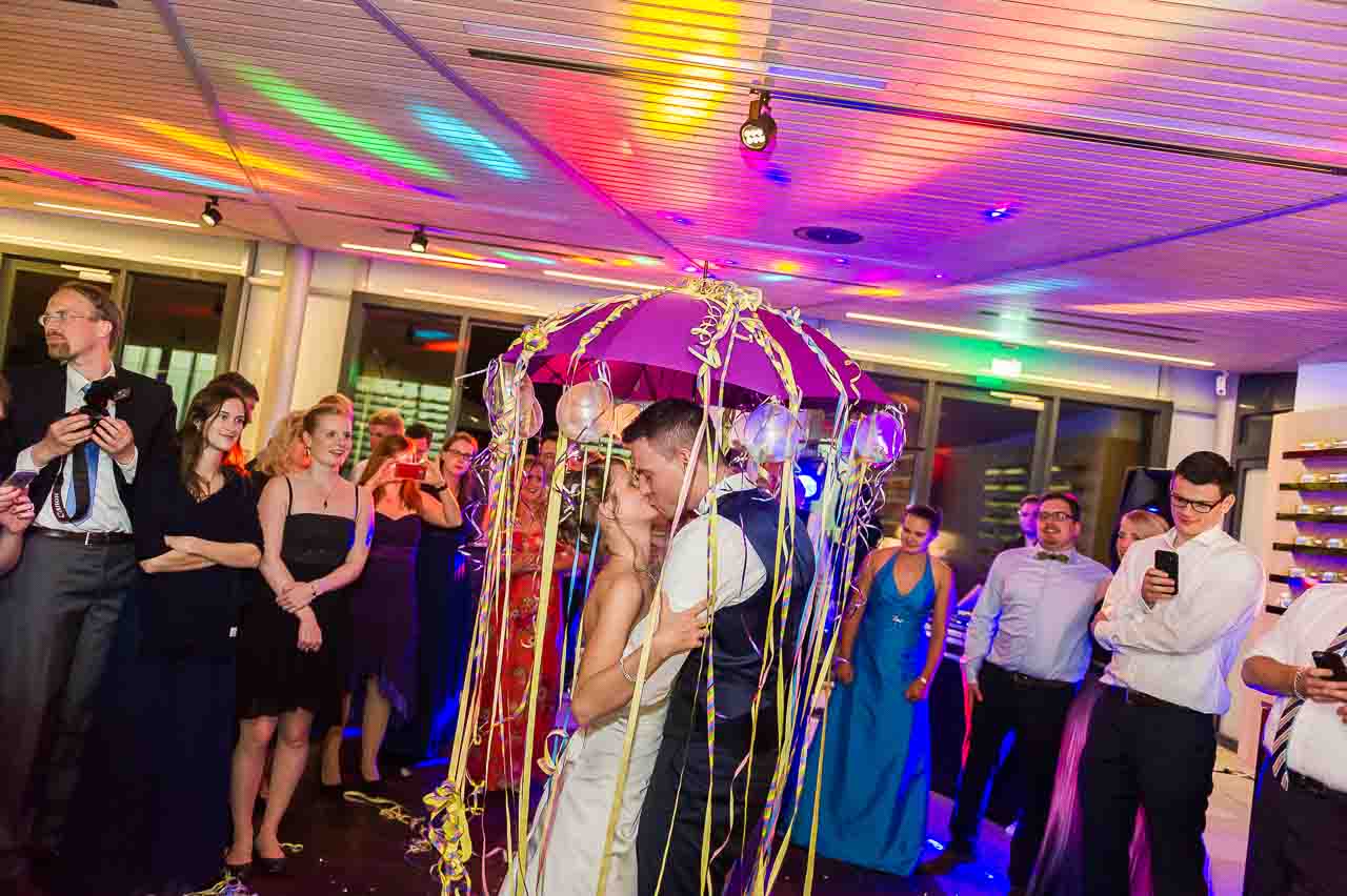 Ein verliebtest Brautpaar kuesst sich innig unter einem festlichen Hochzeitsbaldachin, umgeben von Zuschauern auf ihrer schoenen Hochzeitsfeier.