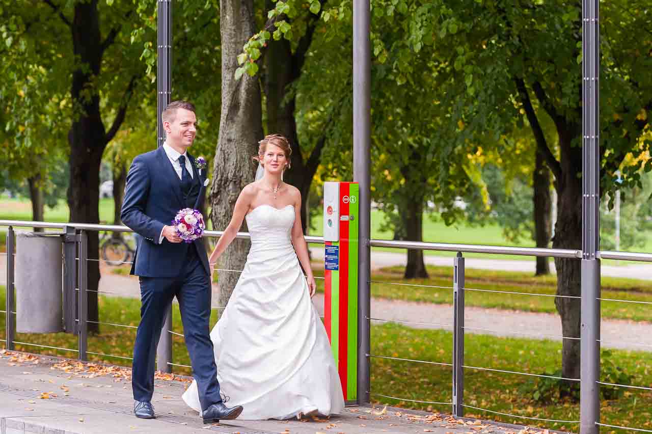 Ein strahlendes Brautpaar spaziert Hand in Hand im Park. Die Braut, in Weiss, laechelt, waehrend der Braeutigam seinen bunten Brautstrauss traegt.