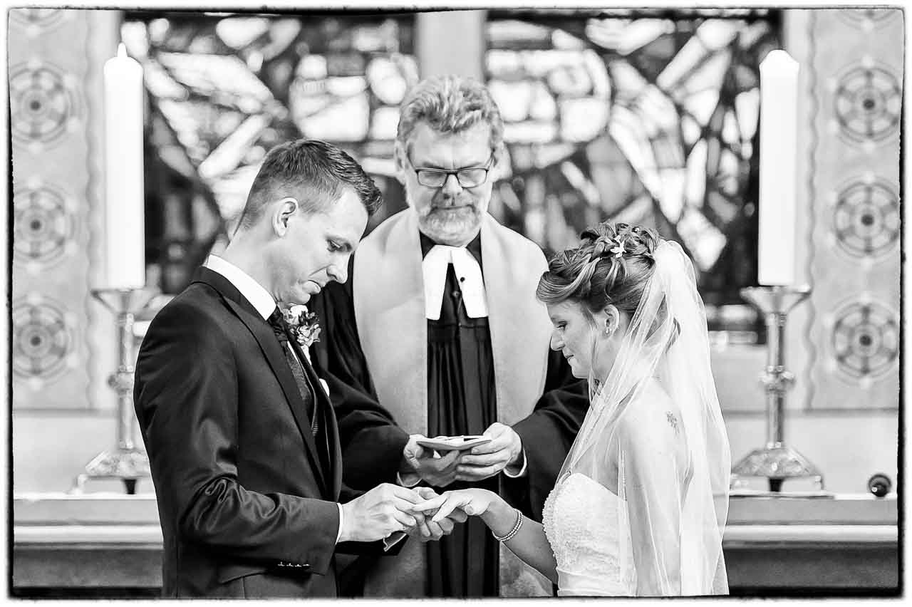 Romantische Schwarzweiss-Aufnahme einer Braut und eines Braeutigams beim Ringwechsel in der Kirche. Ein Pfarrer assistiert dem Paar bei der Zeremonie.