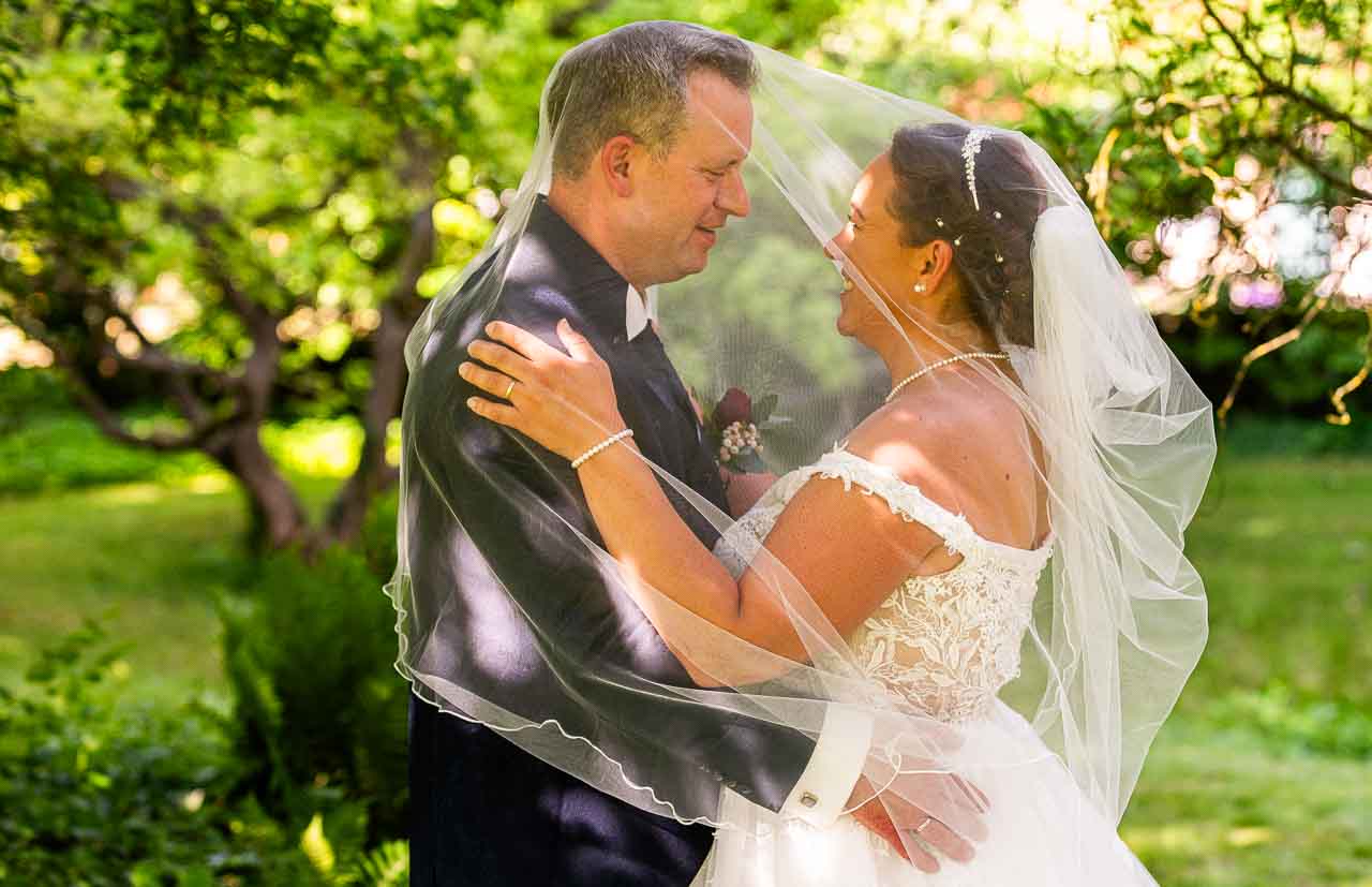 Brautpaar unter Schleier im Gruenen. Die Braut im eleganten, weissen Kleid, der Braeutigam im dunklen Anzug. Liebevoller Blickwechsel im Park.