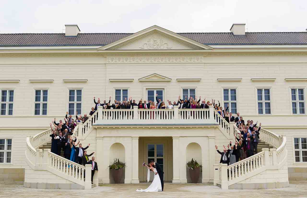 Ein strahlendes Brautpaar kuesst sich vor einem Schloss, umgeben von jubelnden Gaesten auf der Treppe. Eine wunderschoene Hochzeit im Schlossgarten.