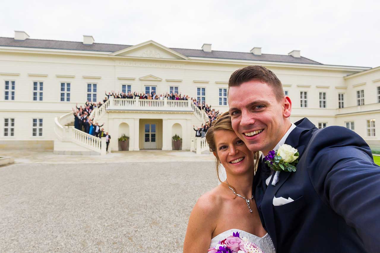 Ein strahlendes Brautpaar macht ein Selfie vor dem beeindruckenden Schloss Herrenhausen. Die Hochzeitsgesellschaft jubelt auf der Treppe im Hintergrund.
