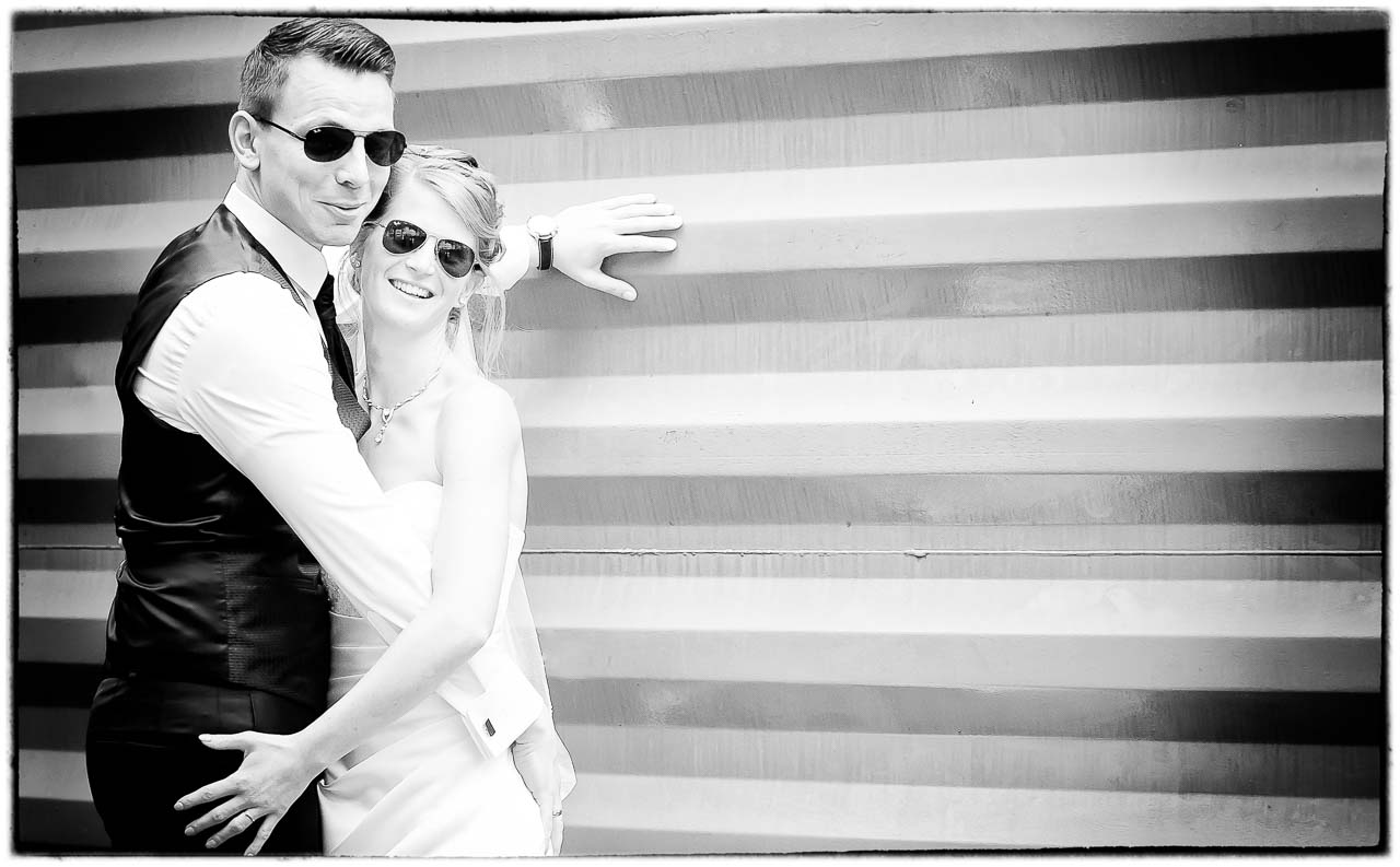 Stilvolles Schwarz-Weiss-Foto eines Braeutigamspaares mit Sonnenbrillen, das sich an eine Wand kuschelt. Die Atmosphaere ist locker und modern.