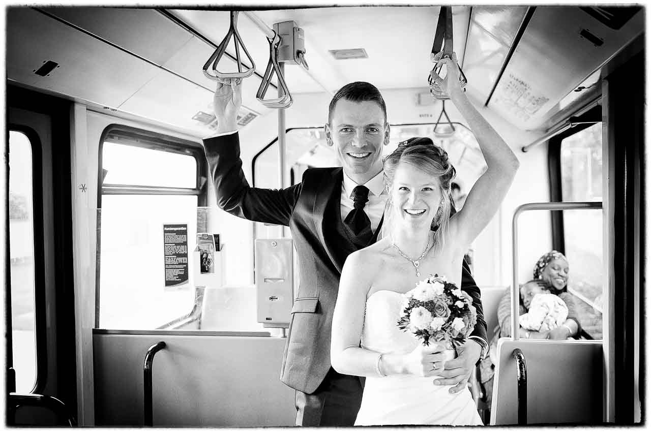 Stilvolles Brautpaar, aufgenommen in einer Strassenbahn. Die Braut strahlt mit ihrem Blumenstrauss, waehrend der Braeutigam elegant laechelt.