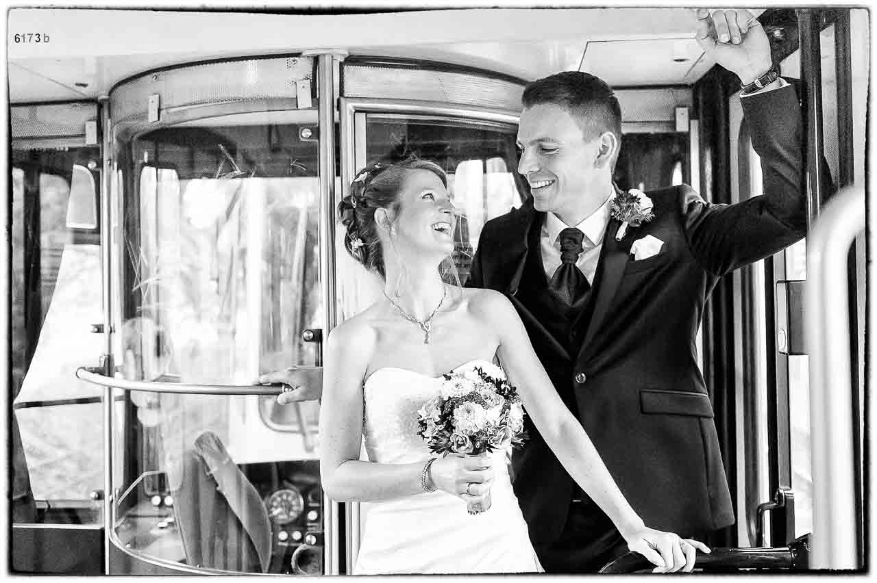 Ein strahlendes Brautpaar, aufgenommen in einer historischen Strassenbahn, blickt sich verliebt an. Die Braut haelt einen zarten Blumenstrauss.