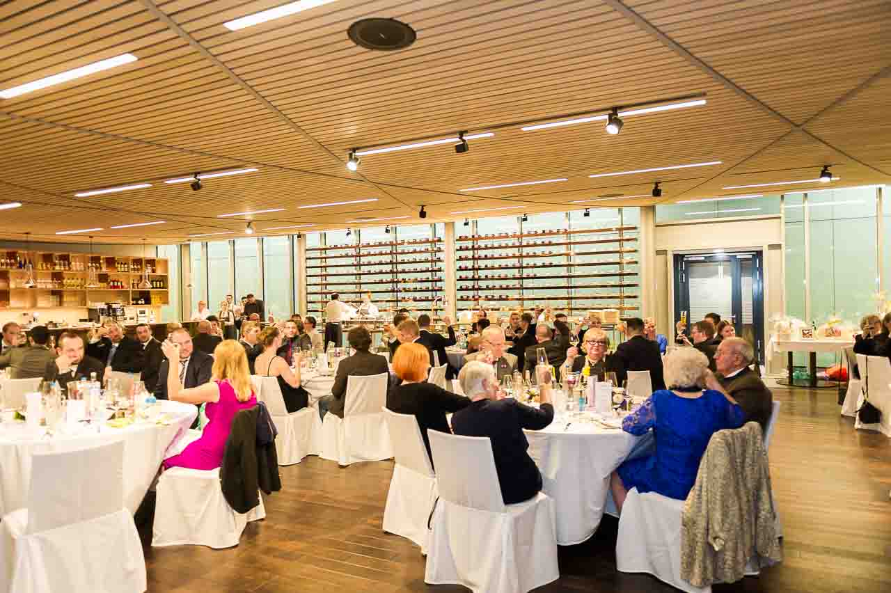 Festliche Gesellschaft in einem eleganten Saal: Eine Gruppe von Menschen sitzt an runden Tischen, gekleidet fuer einen besonderen Anlass oder eine Feier.
