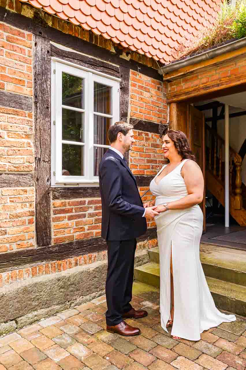 Brautpaar hält Hände in Mosterei Ockensen. Die Braut im eleganten, weißen Kleid, der Bräutigam im dunklen Anzug. Liebevolle Hochzeit.