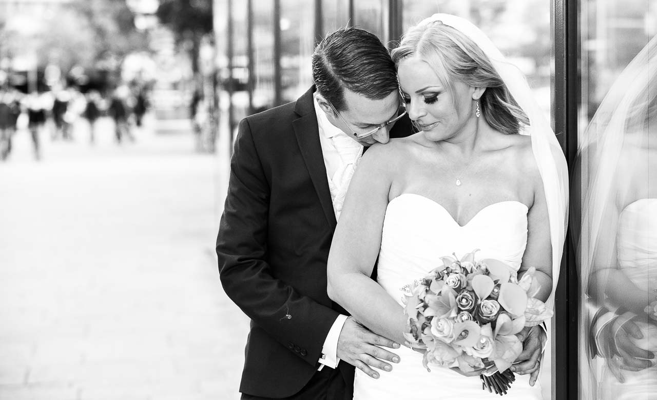 Elegantes Schwarz-Weiss-Foto eines Braeutigams, der seiner Braut zarte Zuneigung schenkt. Sie traegt ein reizendes, schulterfreies Brautkleid und einen Schleier.