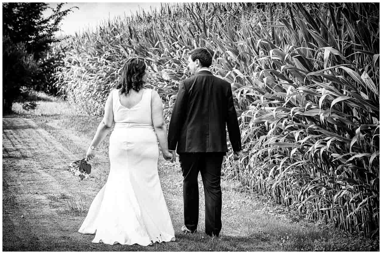 Romantische Hochzeit in Hannover: Brautpaar Hand in Hand am Feldweg. Elegantes Brautkleid, liebevoller Blick. Schwarz-Weiß-Foto voller Zuneigung.