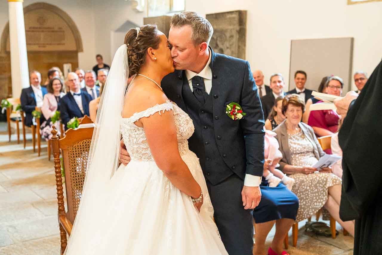 Kirchliche Hochzeit: Brautpaar im innigen Kuss vor den Augen der Hochzeitsgesellschaft. Das Kleid der Braut ist ein echter Hingucker.