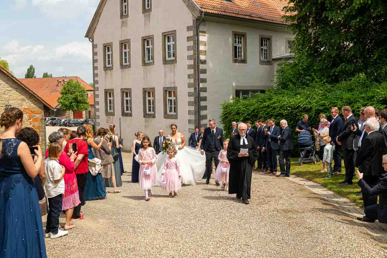 Hochzeitsgesellschaft vor historischem Gebaeude: Braut im weissen Kleid, begleitet von Braeutigam und Blumenmaedchen, auf dem Weg zur Trauung.