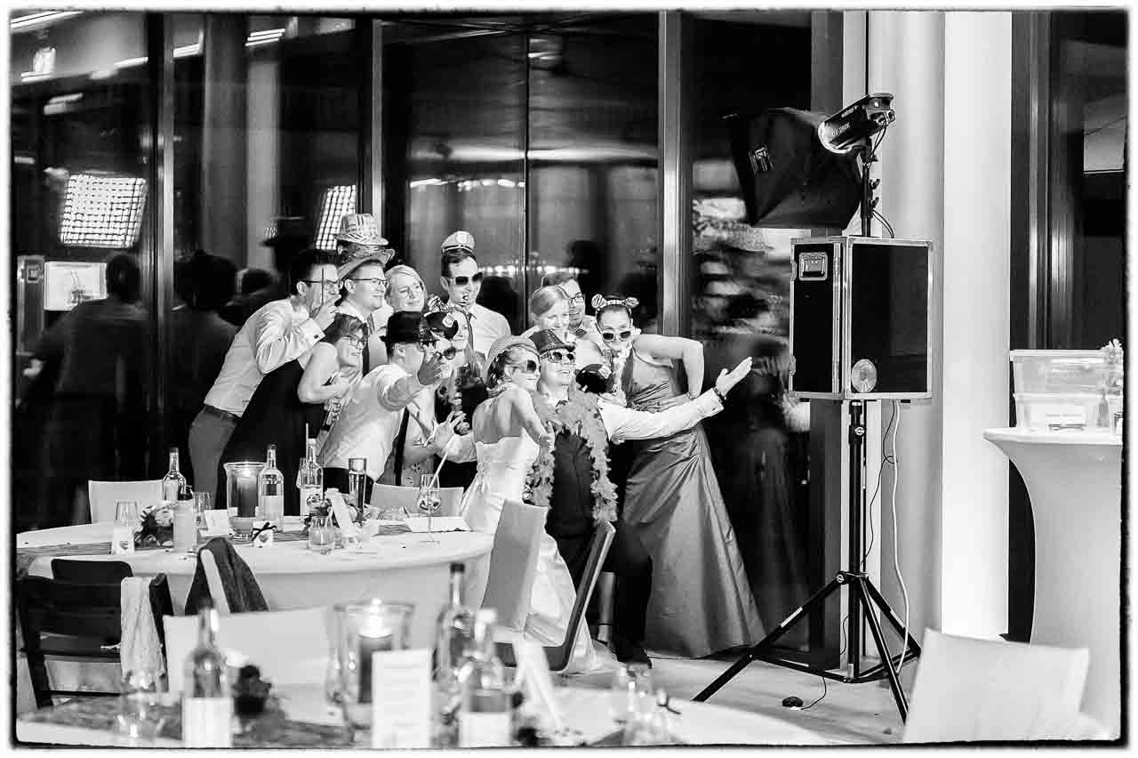 Eine festliche Hochzeitsgesellschaft posiert ausgelassen vor einer Fotobox. Die Braut strahlt, waehrend die Gaeste mit Accessoires fuer lustige Erinnerungen sorgen.