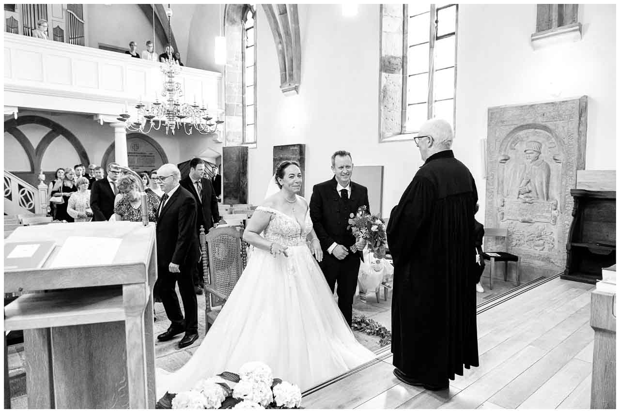 Kirchliche Hochzeit: Braut im eleganten Kleid, Braeutigam und Pfarrer in der Kirche. Festlicher Moment voller Liebe und Tradition.