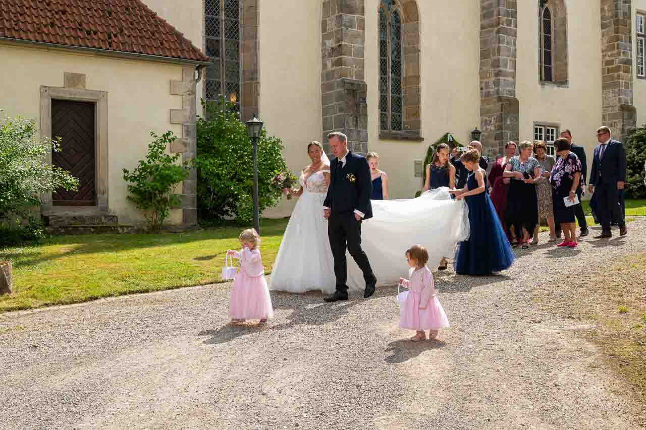 Romantische Hochzeit vor der Kirche: Brautpaar mit Blumenmaedchen und Gaesten. Das lange, weisse Kleid der Braut ist ein echter Hingucker.