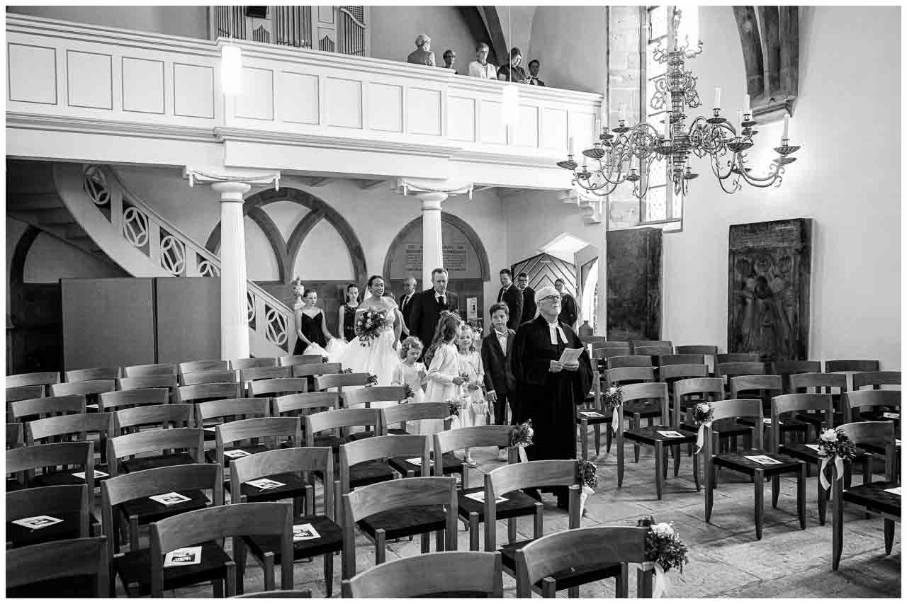 Emotionale Hochzeitszeremonie in der Kirche: Die Braut schreitet mit ihrem Vater zum Altar, begleitet von Blumenmaedchen und Braeutigam.