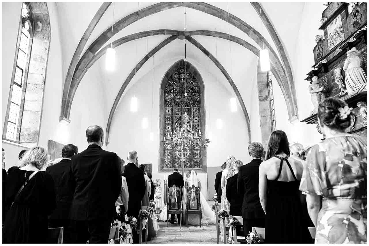 Feierliche Hochzeitszeremonie in der Kirche: Brautpaar vor Altar, umgeben von Gästen. Elegante Architektur und festliche Atmosphaere in Schwarz-Weiss.