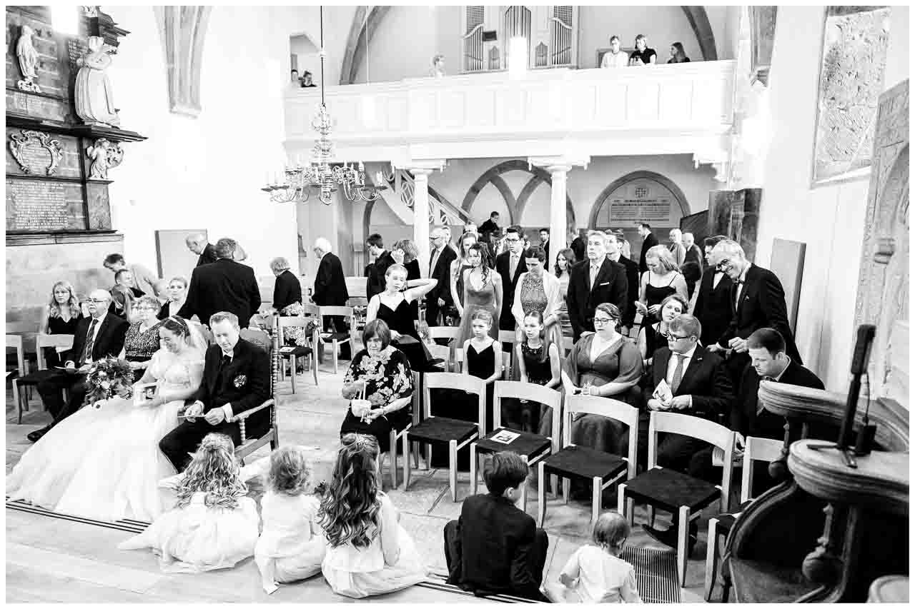 Hochzeitsgesellschaft in der Kirche: Brautpaar umgeben von Gaesten, Blumenmaedchen sitzen am Gang. Festliche Atmosphaere in Schwarz-Weiss.