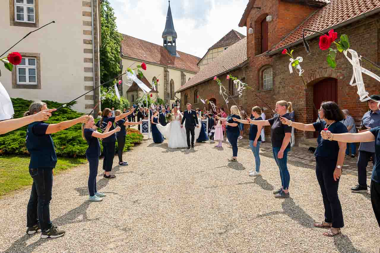 Hochzeitspaar verlaesst die Kirche, Spalier mit Rosen geschmueckt. Braut in Weiss, Braeutigam im Anzug. Romantische Atmosphaere nach der Trauung.