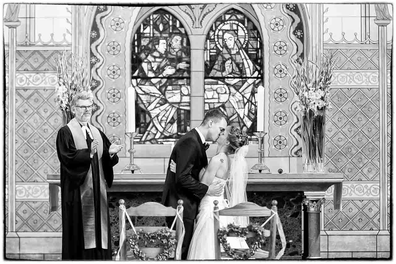Romantischer Hochzeitskuss in der Kirche: Ein Brautpaar teilt einen innigen Moment vor einem beeindruckenden Glasfenster. Schoen und unvergesslich!
