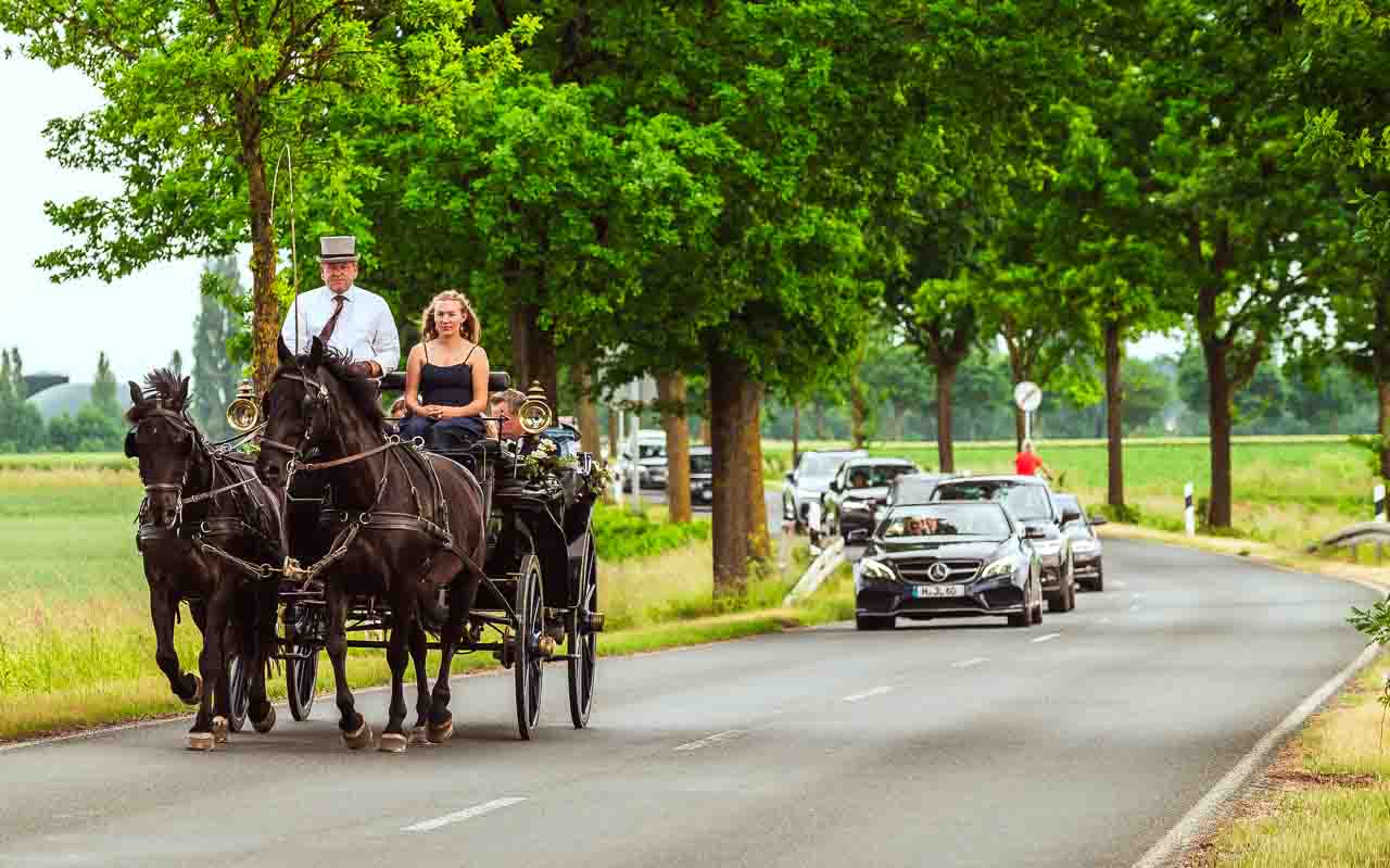 Romantische Hochzeitskutsche mit Brautpaar auf dem Land. Schwarze Pferde ziehen die Kutsche auf einer Allee entlang, ein maerchenhaftes Erlebnis.