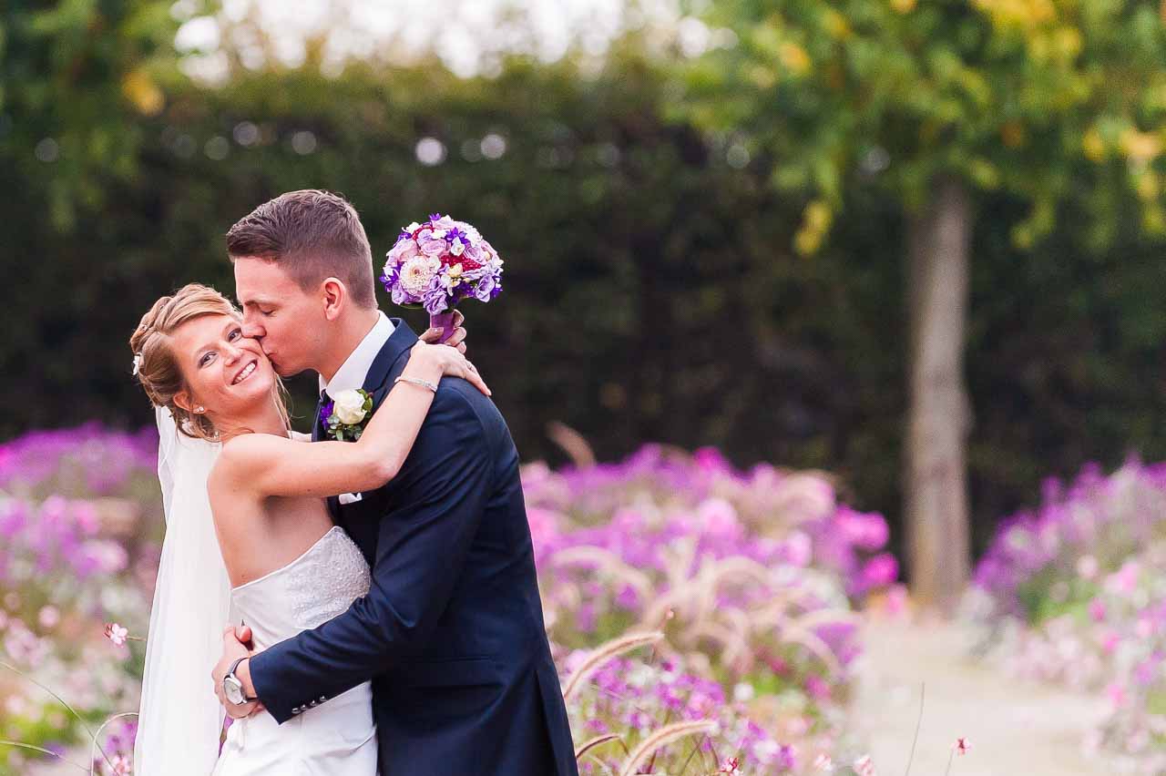 Ein zaertlicher Hochzeitsmoment im Park: Der Braeutigam gibt seiner strahlenden Braut einen Kuss. Ein schoenes Brautpaar inmitten bluehender Blumen.