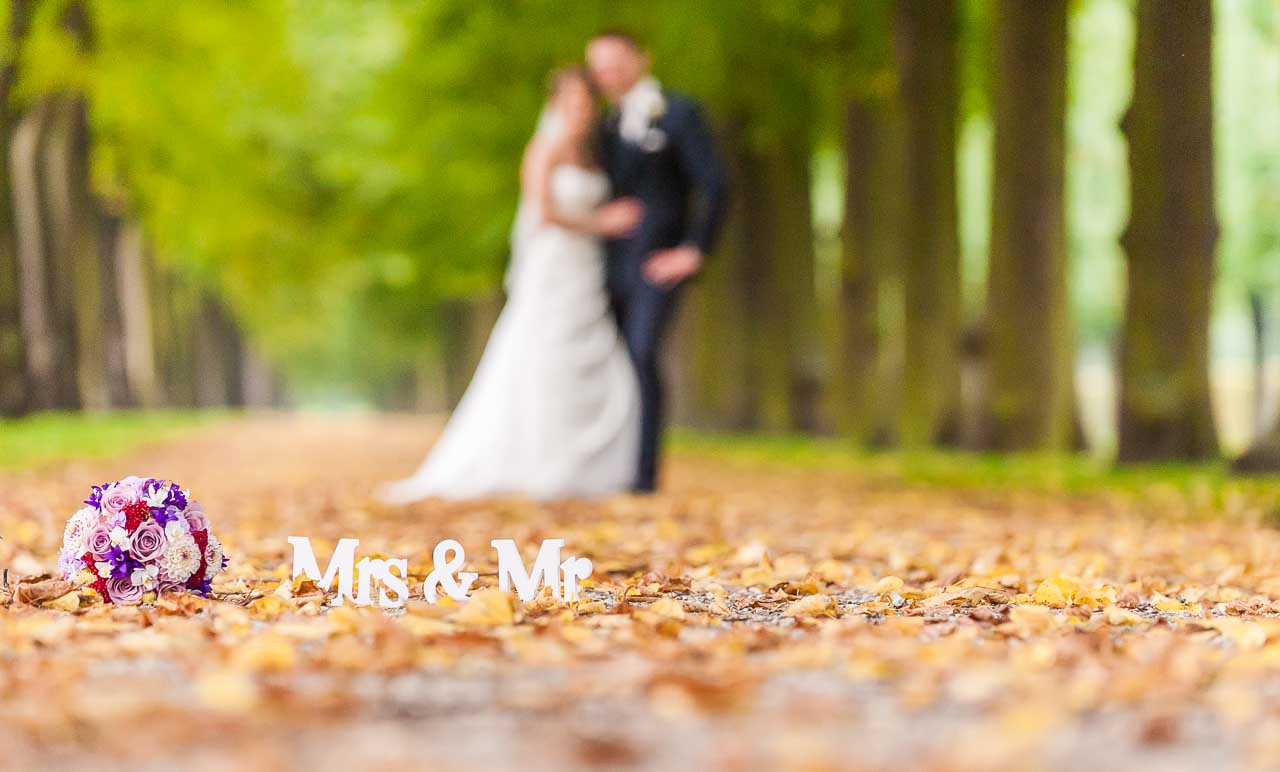 Romantische Hochzeit im Park: Ein Brautstrauss und die Worte 'Mrs & Mr' liegen auf dem herbstlichen Laub. Im Hintergrund verschwommen das Brautpaar.