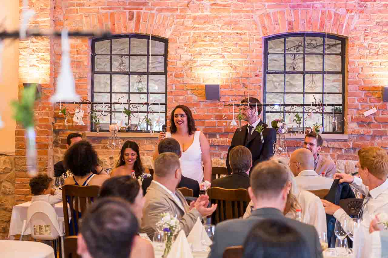 Hochzeit in der Mosterei Ockensen: Braut hält Rede vor rustikalem Backstein-Hintergrund. Liebevolle Details, festliche Atmosphäre.