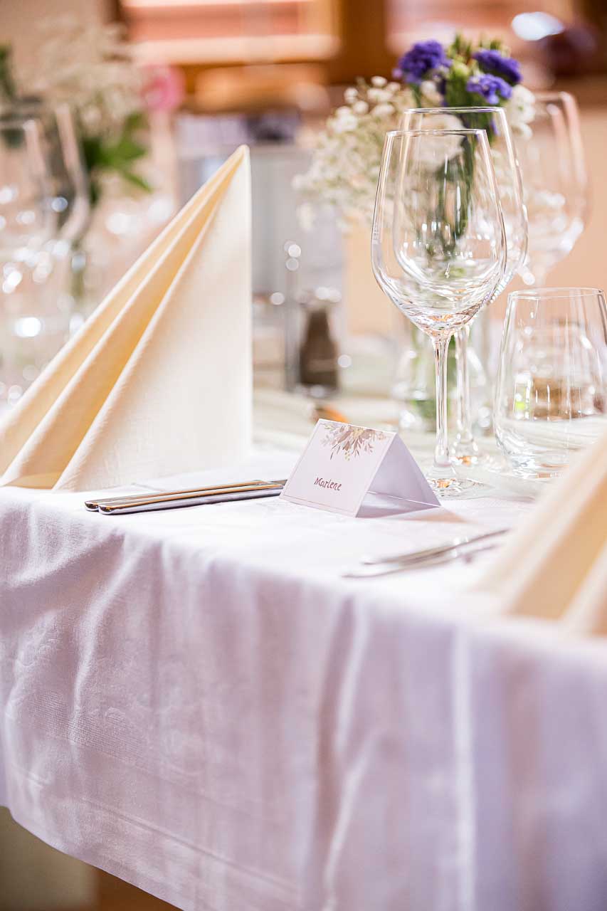 Festliche Hochzeitstafel in Ockensen: Elegante Gläser, zarte Blumenarrangements und eine persönliche Namenskarte für Marlene. Stilvolle Tischdeko.