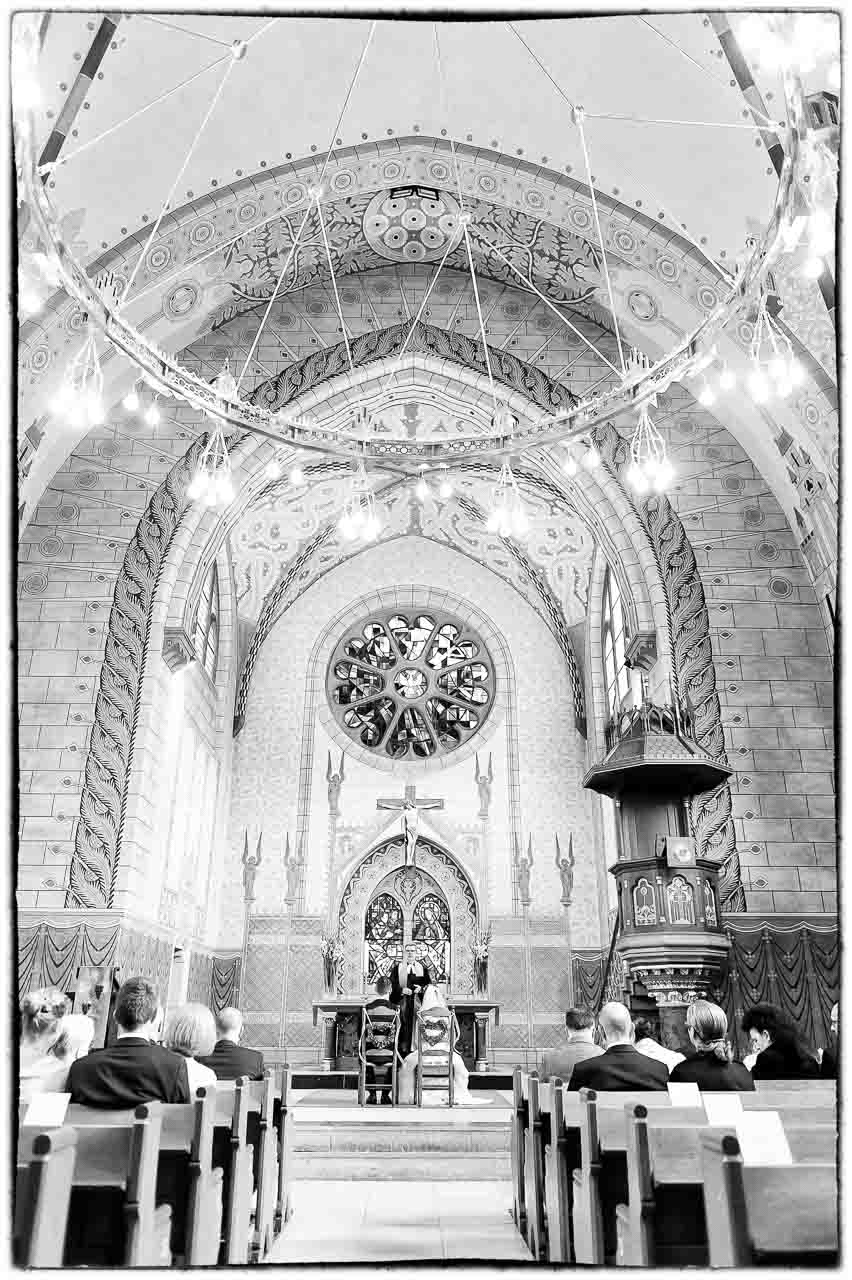 Elegante schwarz-weisse Aufnahme einer Trauung in der Kirche. Das Brautpaar steht vor dem Altar, umgeben von sakraler Architektur und sanftem Licht.