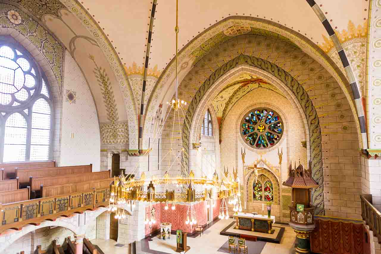 Wunderschoene Innenansicht einer Kirche mit kunstvollen Malereien und einem grossen, bunten Glasfenster. Elegante Details und warme Farben laden zum Traeumen ein.
