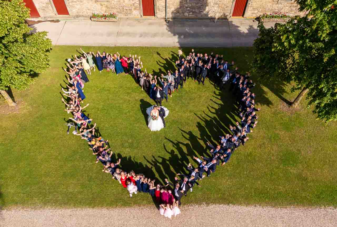 Luftaufnahme: Brautpaar und Hochzeitsgesellschaft formen ein Herz auf gruener Wiese. Romantische Szene mit liebevollen Details.