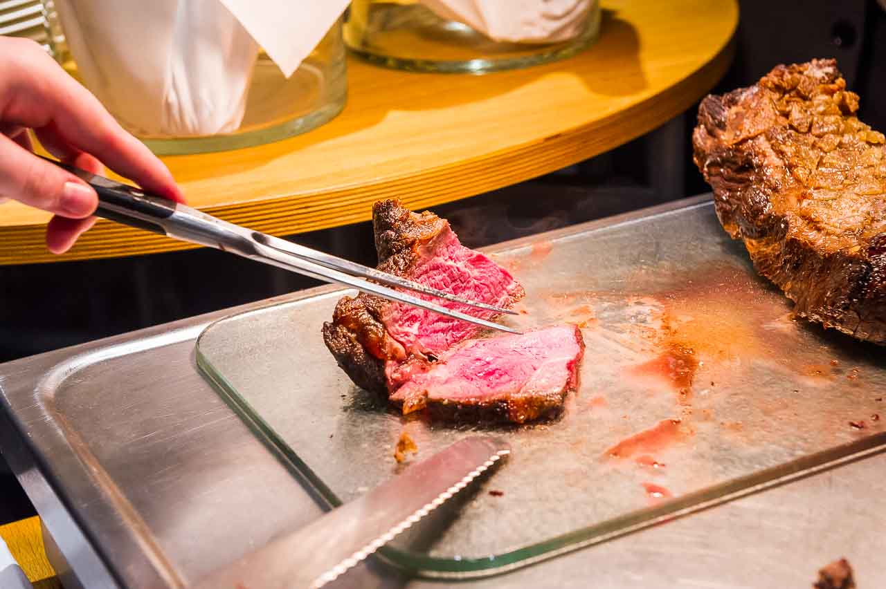 Ein zartes, rosa gebratenes Stueck Fleisch wird mit einer Zange gehalten. Die Textur und der Saftigkeit des Fleisches laden zum Genuss ein. Ein Fest fuer Feinschmecker!