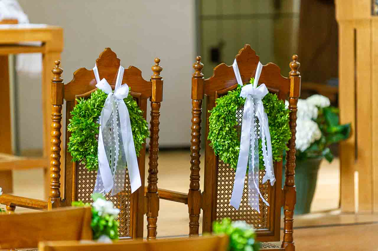 Dekorierte Stuehle in der Kirche mit gruenen Kraenzen und weissen Schleifen. Romantische Hochzeitsdekoration fuer eine stilvolle Trauung.