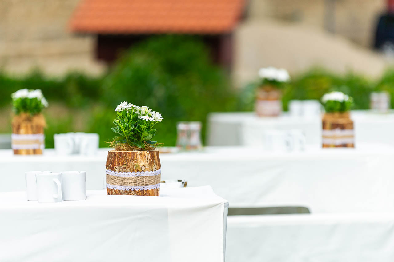 Romantische Tischdeko mit Blumen in Holztoepfen. Zarte, weisse Blueten und rustikales Holz fuer eine stilvolle Hochzeitsfeier im Freien.