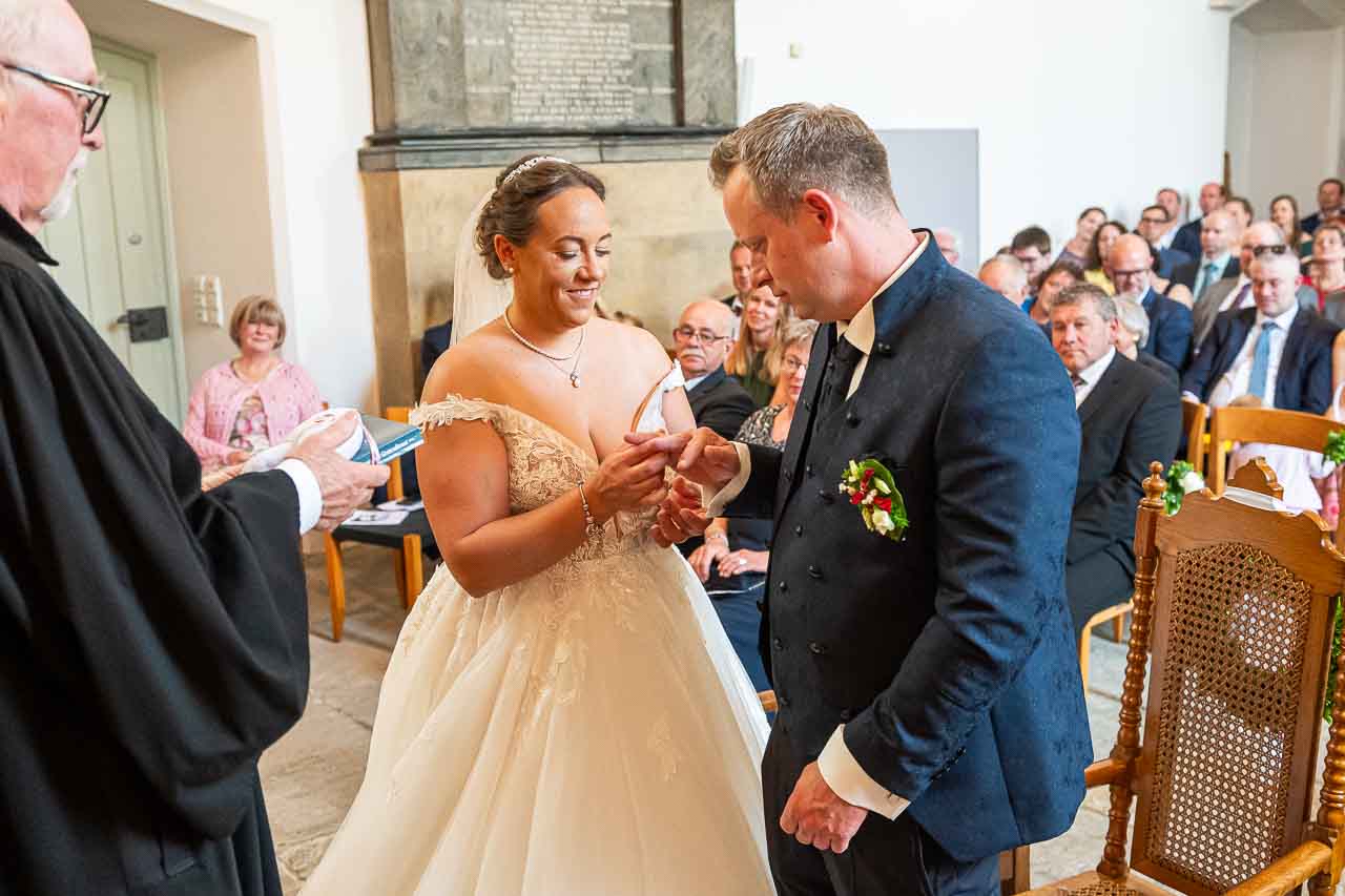 Romantische Trauung in der Kirche: Braeutigam steckt der strahlenden Braut den Ring an. Festliche Atmosphaere, liebevolle Details.